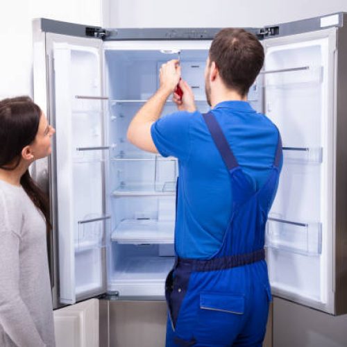 Woman Looking At Male Repairman Fixing Refrigerator With Screwdriver